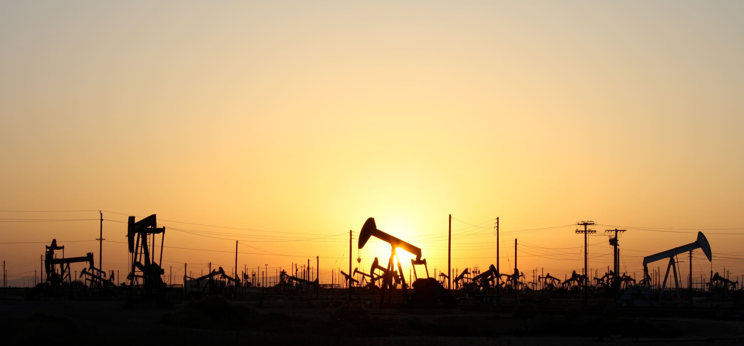 Silhouetted oil pumpjacks across a flat oil field against a glowing orange sunset sky